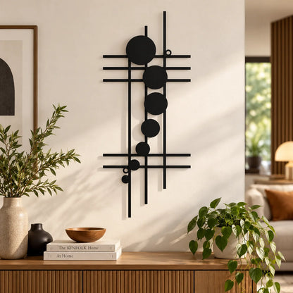 A black metal abstract wall art piece with vertical and horizontal lines and circular elements, displayed on a white wall in a modern living room setting above a wooden console table with plants and books.