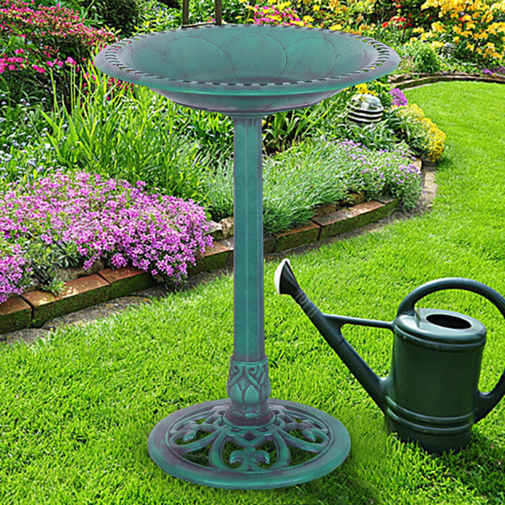 A teal metal garden pedestal bird bath stands on a green lawn next to a dark green watering can, with a colorful flower garden in the background.