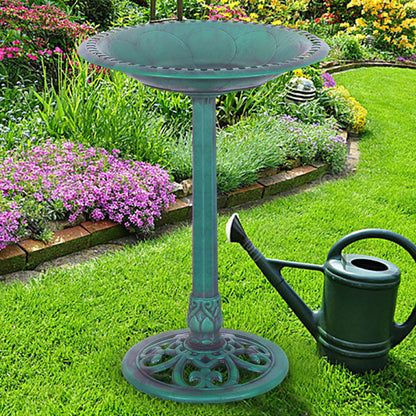 A teal metal garden pedestal bird bath stands on a green lawn next to a dark green watering can, with a colorful flower garden in the background.